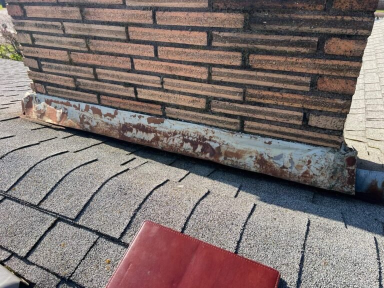 Brick chimney base with heavily rusted metal flashing where it meets the old shingle roof.