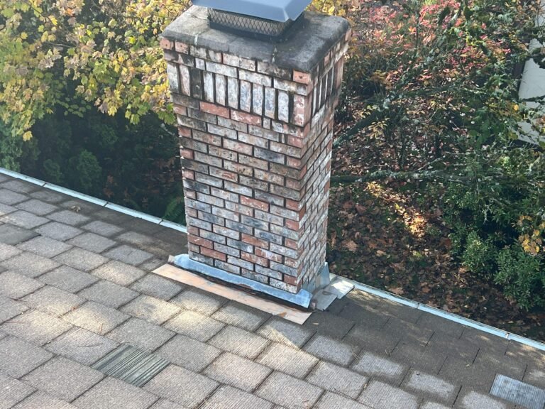 Brick chimney with metal flashing rising from a weathered gray shingle roof, surrounded by autumn foliage.