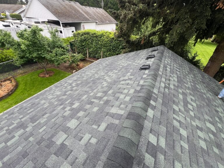New gray and green-toned shingle roof on an outbuilding, with box vents near the ridge and a backyard with trees and a neighboring house.