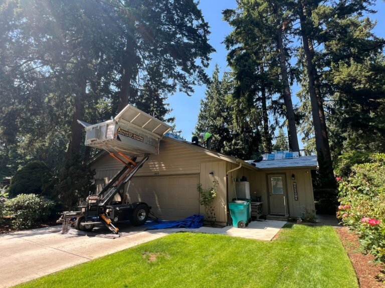 Roofing conveyor lift tilted up against the roof of a tan single-story house, with a debris trailer and green trash bin below.