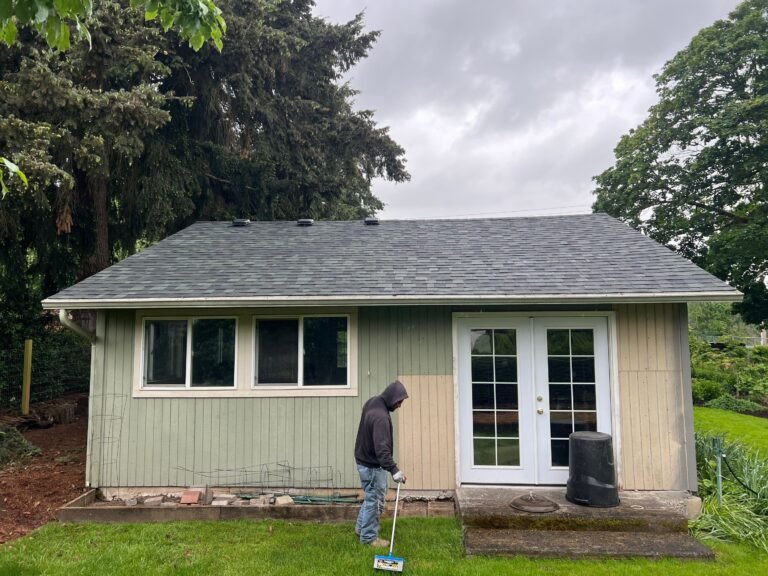 Sage green garden building with white double doors and a new gray shingle roof, with a worker in a hooded jacket using a magnetic sweeper on the lawn.