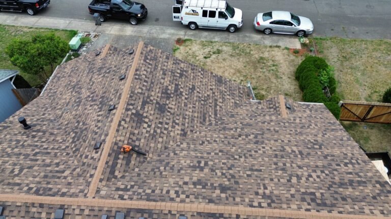 Aerial view of a new brown shingled roof with ridge caps and a leaf blower resting on the surface, above a driveway with a van and cars.