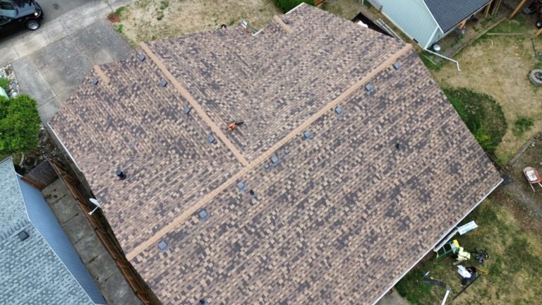 Another aerial angle of the finished brown shingle roof, showing hip and ridge lines and vents, above a yard and neighboring rooftops.