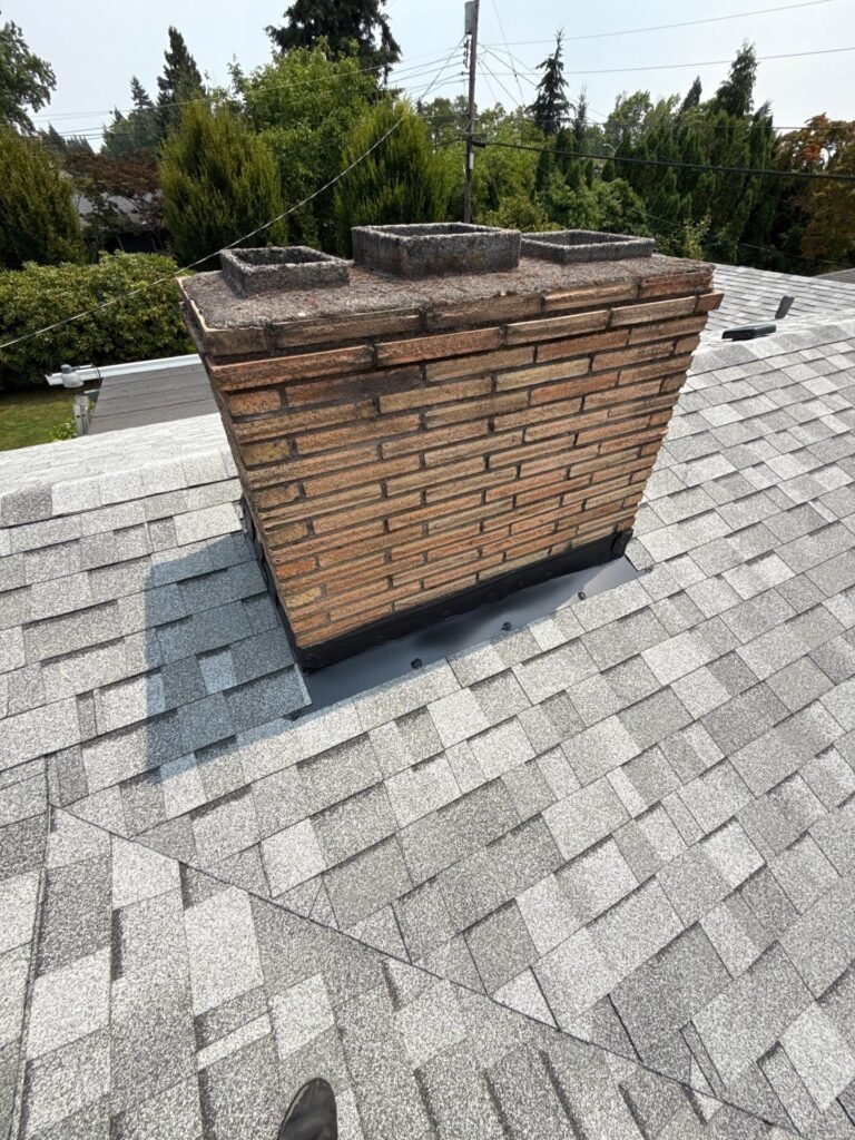 Brick chimney with new light gray shingles and fresh black flashing installed around its base.