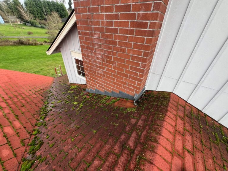 Red brick chimney rising from an old red shingle roof covered in moss, with white board-and-batten siding beside it.