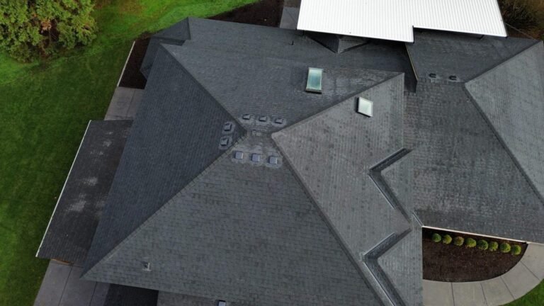 Top-down view of the finished dark gray shingle roof with skylights and vents, over a landscaped yard.