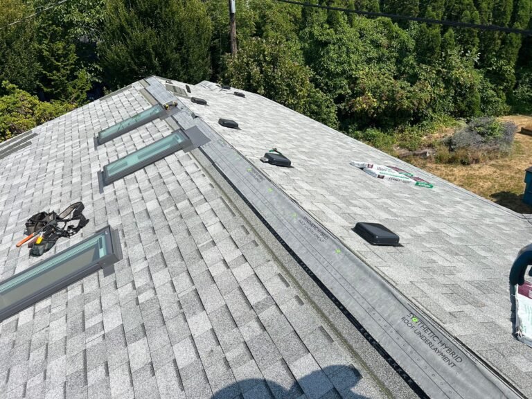 Roof in progress with skylights, black box vents, and a ridge section partly covered, with tools and shingle bundles on the surface and trees behind.