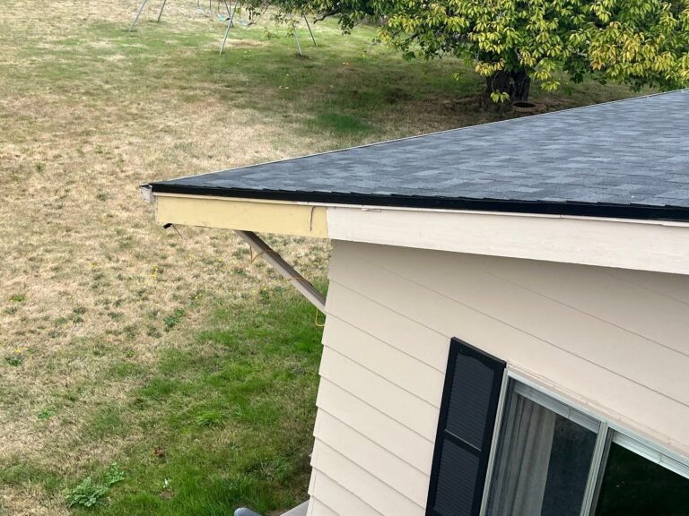 Roof corner at the gable end, showing clean shingle edges and drip edge above the tan siding and a dark-framed window.