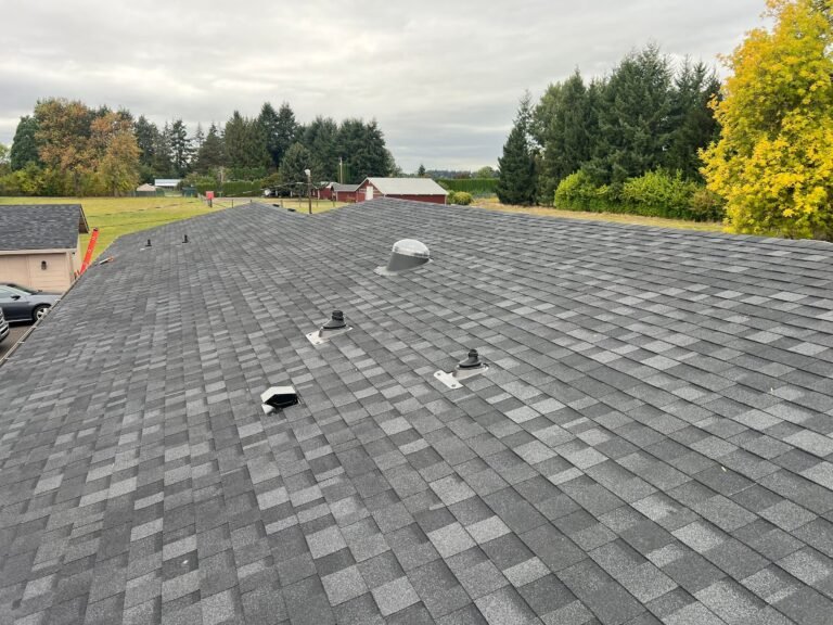 New gray asphalt shingles covering a roof slope, with several vent pipes and a round roof vent, and a red barn and trees in the distance.