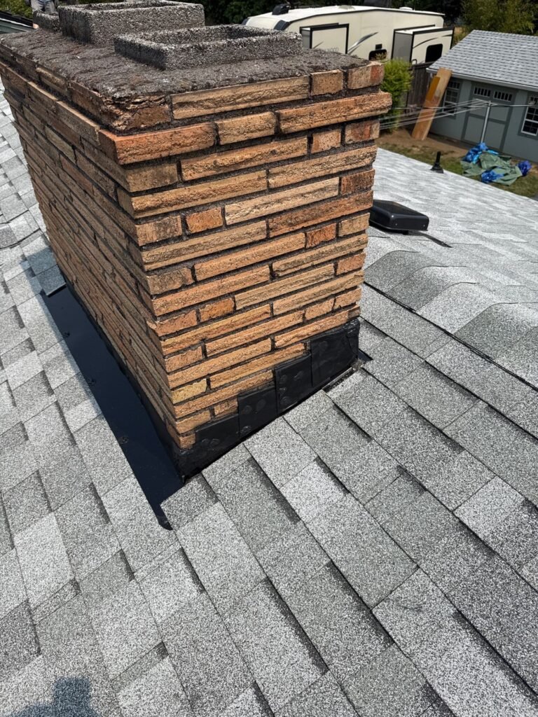 Chimney from another angle, with black flashing partly installed at the base and a box vent on the new shingles nearby.