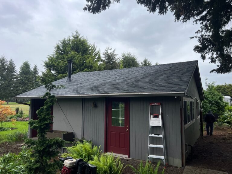 Gray board-and-batten outbuilding with a red door, a stepladder by the entry, and a new dark gray shingle roof.