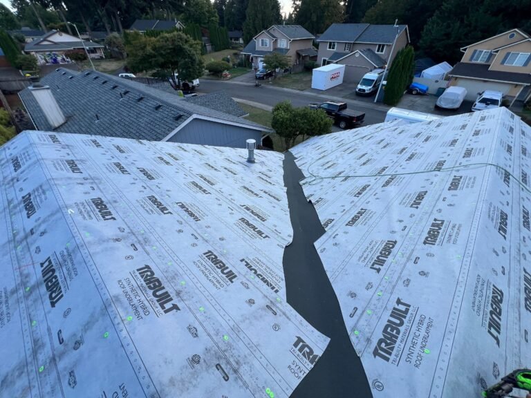 Roof ridge and valley covered in synthetic underlayment, with a dark waterproof membrane running down the valley and a suburban street beyond.