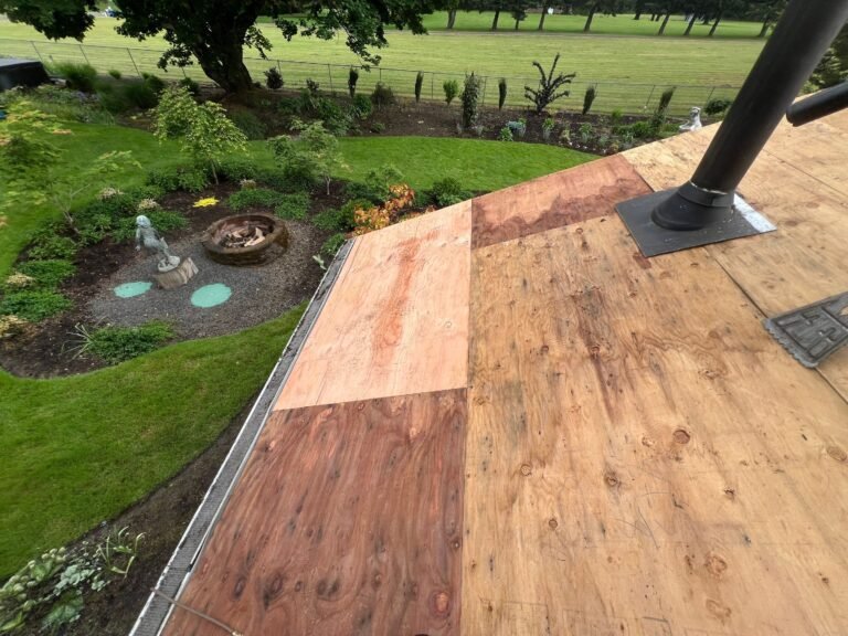 Roof decking with new and replaced plywood panels around a black vent pipe, above a landscaped garden with a fire pit and a green lawn.