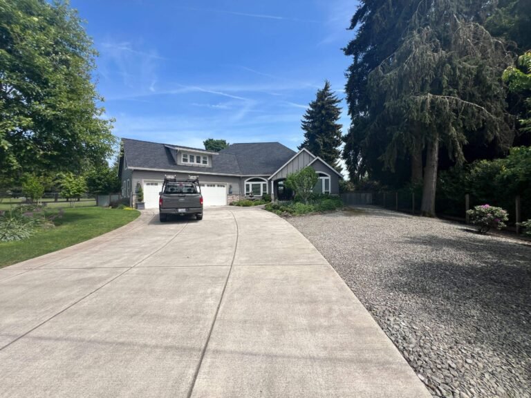 Wide concrete driveway leading to a gray home with a three-car garage and a pickup truck parked in front, surrounded by tall evergreens.