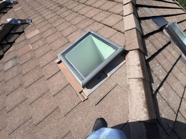Skylight with a copper-toned flashing on an aging brown shingle roof, next to a ridge line.