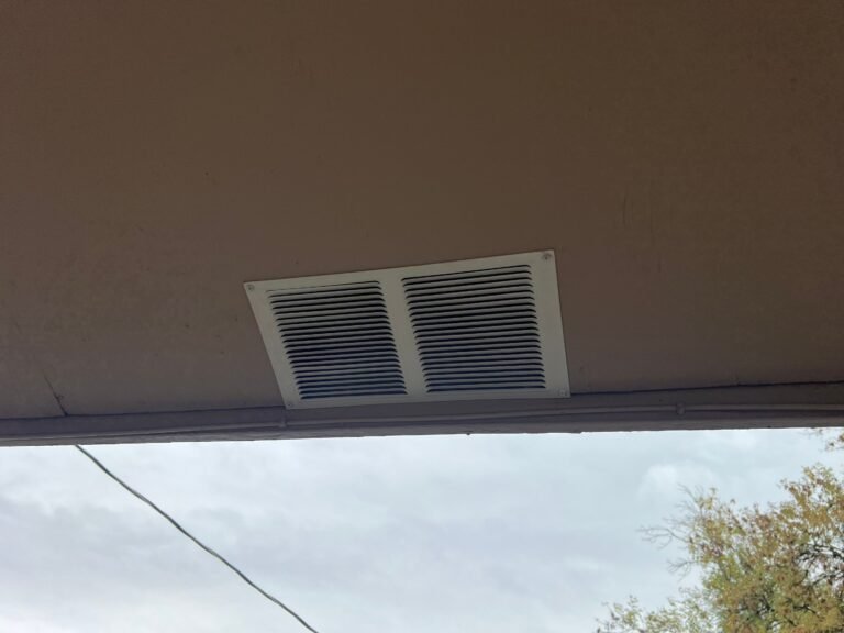 Close-up of a rectangular louvered soffit vent set into the underside of the eave, with a power line and sky beyond.