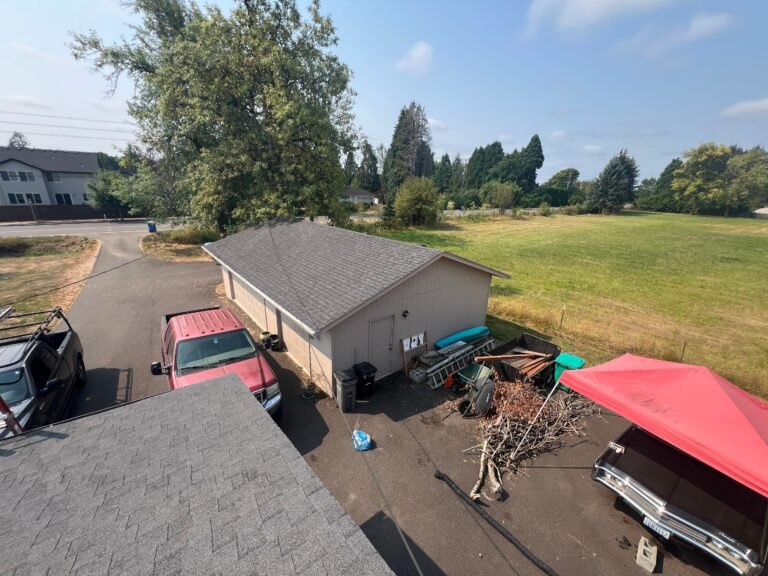 Aerial view of a detached garage with a gray shingled roof beside a gravel driveway, with a red pickup truck and a pile of branches nearby.