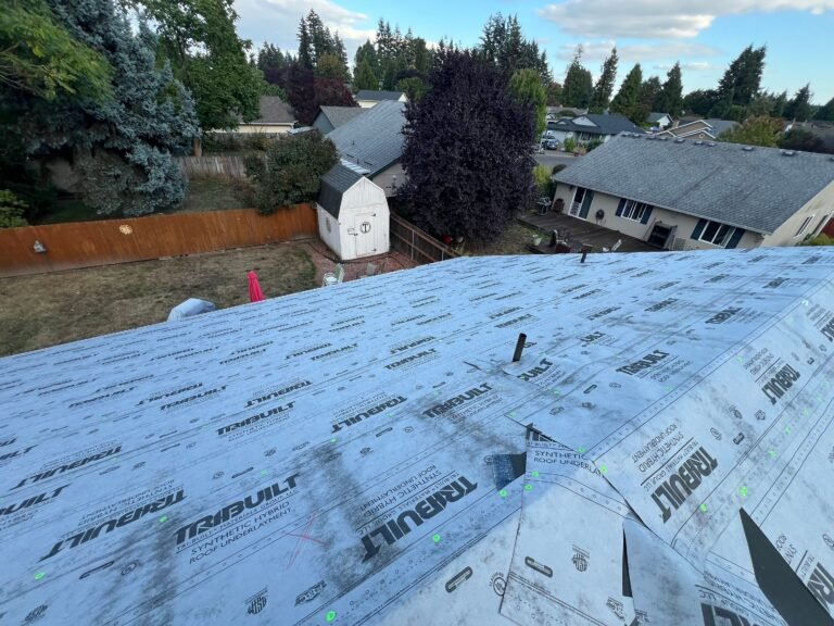 Roof covered in blue-gray synthetic underlayment with a plumbing vent pipe, overlooking a fenced backyard with a shed and neighboring houses.