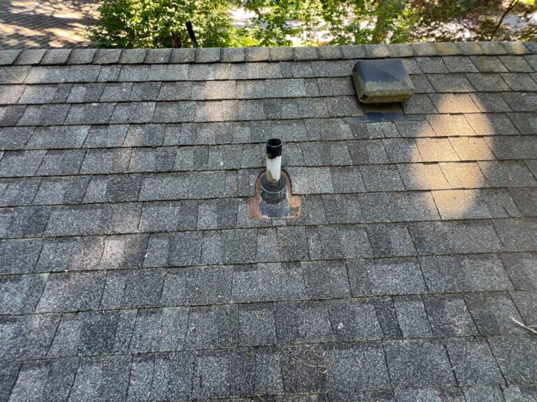 Aged shingle roof slope with a metal plumbing vent pipe and a box vent near the ridge, dappled with tree shadows.
