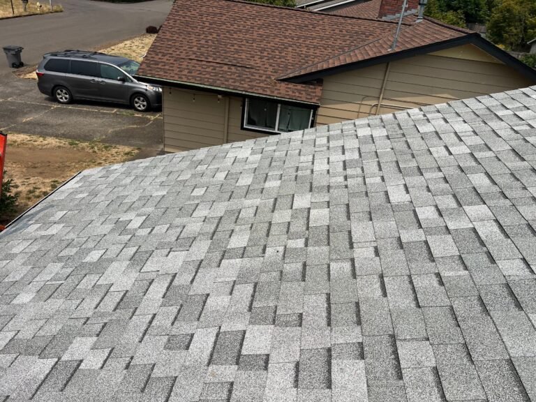 Finished gray shingle roof in the foreground, with a tan house with a brown roof and a minivan parked on the street beyond.