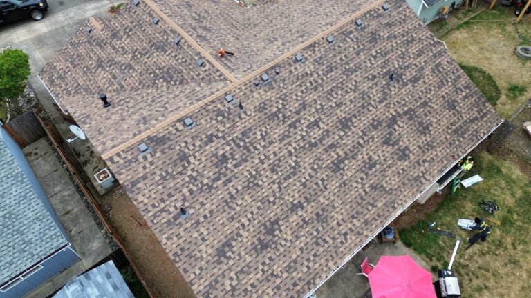 Overhead view of a completed brown shingle roof with several valleys, box vents, and plumbing vents, over a backyard with a pink patio umbrella.