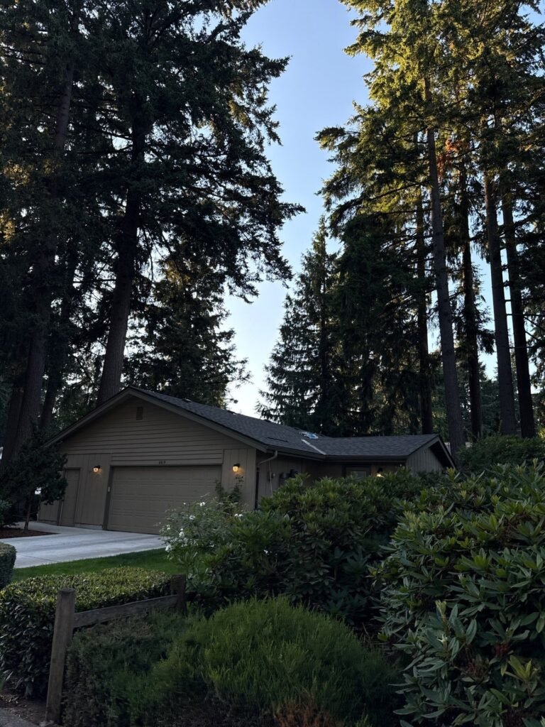 Tan single-story home with a two-car garage and a dark shingle roof, framed by tall evergreen trees and shrubs.