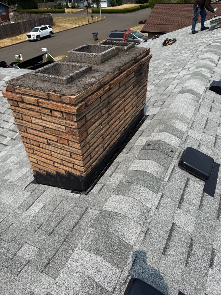 Brick chimney with a concrete cap, surrounded by new gray shingles and finished black flashing, with a vent beside it.