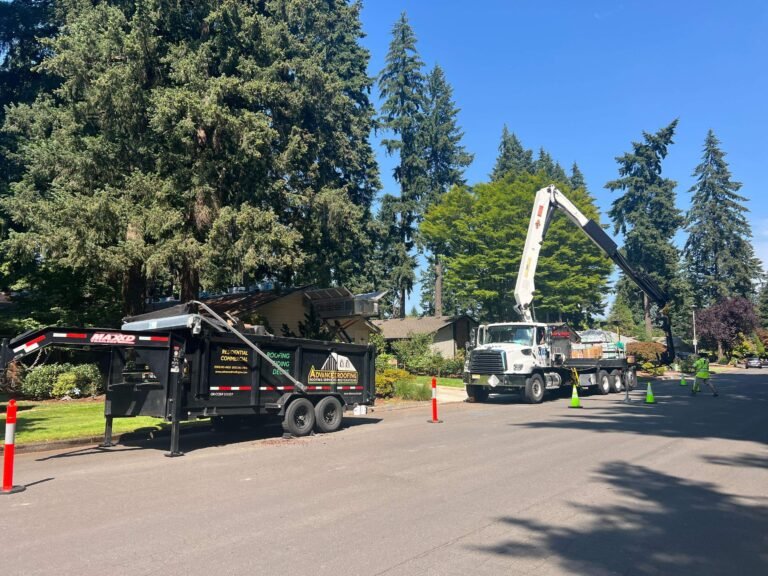 Boom truck and black dump trailer parked on a residential street with safety cones, ready for a roofing job, with tall evergreens behind.