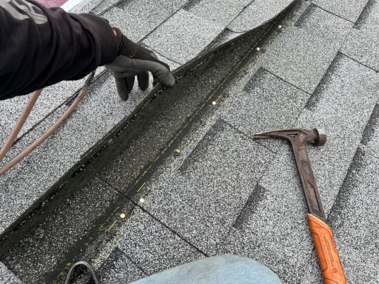 Gloved hand lifting a shingle edge to reveal dark staining underneath, with a hammer lying on the roof.
