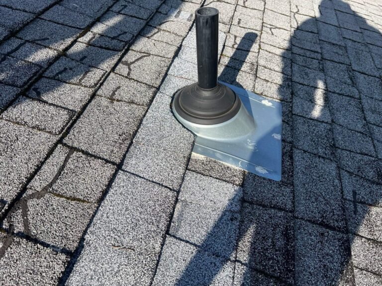 Gray asphalt shingle roof with a black plumbing vent pipe on a metal flashing base, with the shadow of a person cast across the shingles.