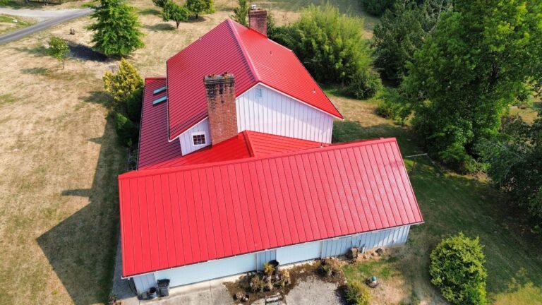 Aerial view of the full red metal roof, showing the garage wing, main gable, and brick chimney above the surrounding field and trees.