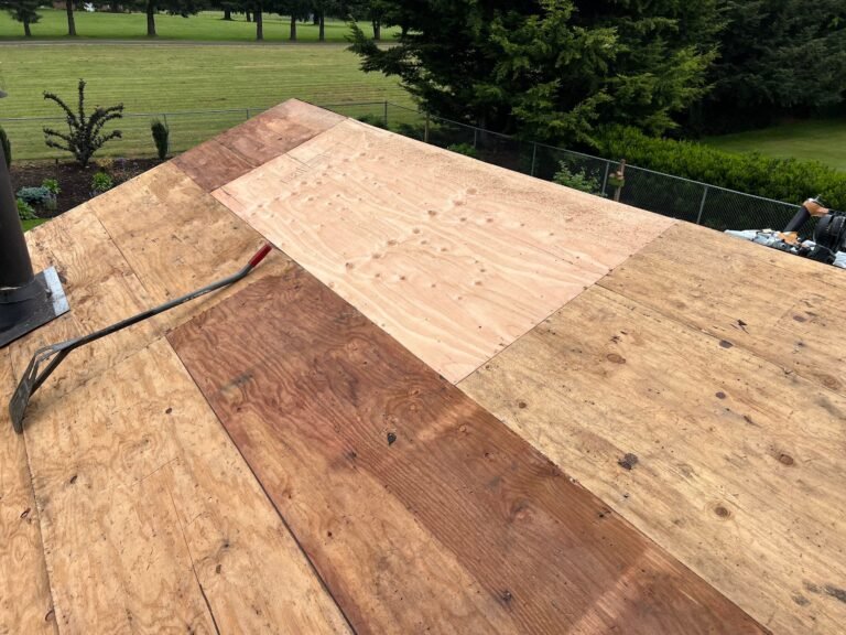 Roof ridge with a mix of new and weathered plywood decking, a roofing tool resting on the surface, and a golf course beyond.