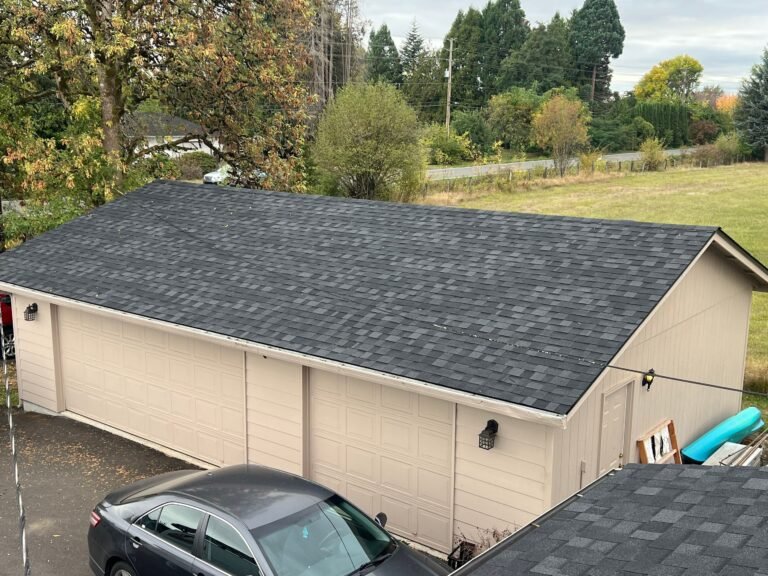 Garage roof with fresh dark gray shingles, seen from above, with a parked car below and trees and fields in the background.
